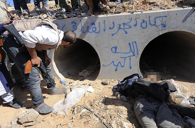 A Libyan National Transitional Council (NTC) fighter looks through a large concrete pipe where ousted Libyan leader Moamer Kadhafi was allegedly captured, with a dead loyalist gunmen in the foreground, in the coastal Libyan city of Sirte on October 20, 2011. A Libyan National Transitional Council (NTC) commander had told AFP that Kadhafi was captured as his hometown Sirte was falling, adding that the ousted strongman was badly wounded. Arabic graffiti in blue reads: "This is the place of Kadhafi, the rat.. God is the greatest.