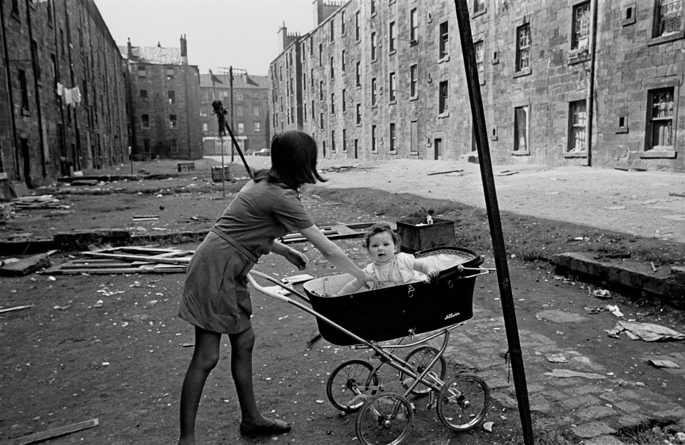  Mother and baby in Gorbals tenement courtyard 1970: A woman pushes her daughter through a filthy street in a pram