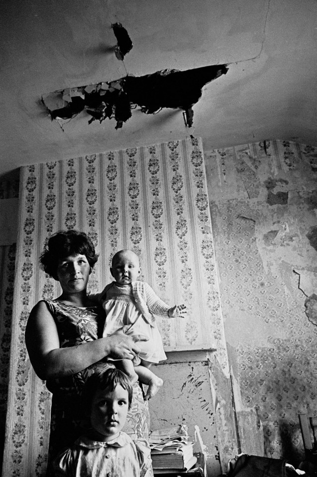  Mother and baby, slum property Sparkbrook, 1971: A mother holds her baby girl and stands with her daughter in an room with a hole in the ceiling