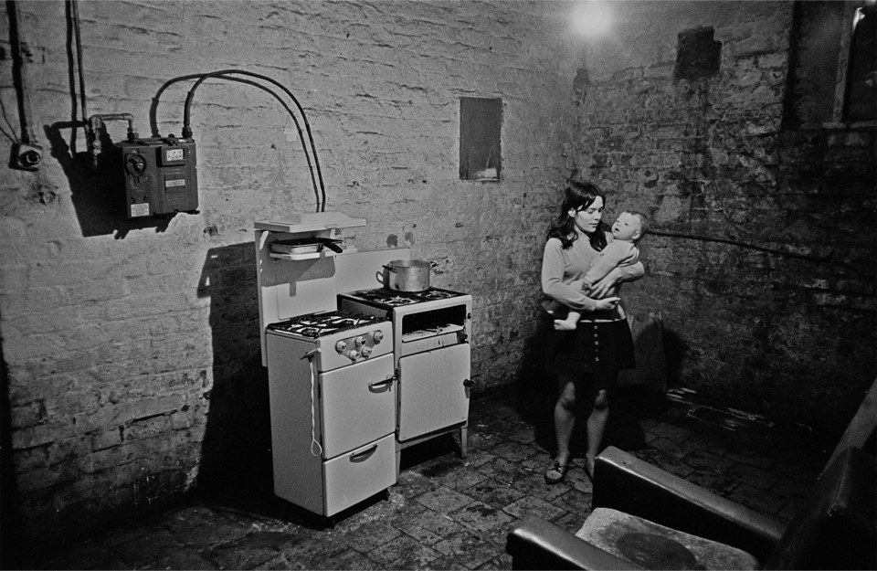  Kitchen in a Liverpool multi-let, 1969. A mother clutches her toddler in a dark room