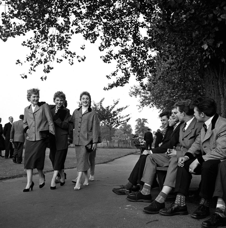  A group of teddy boys sitting on a park bench by a huge tree as they watch girls walking by