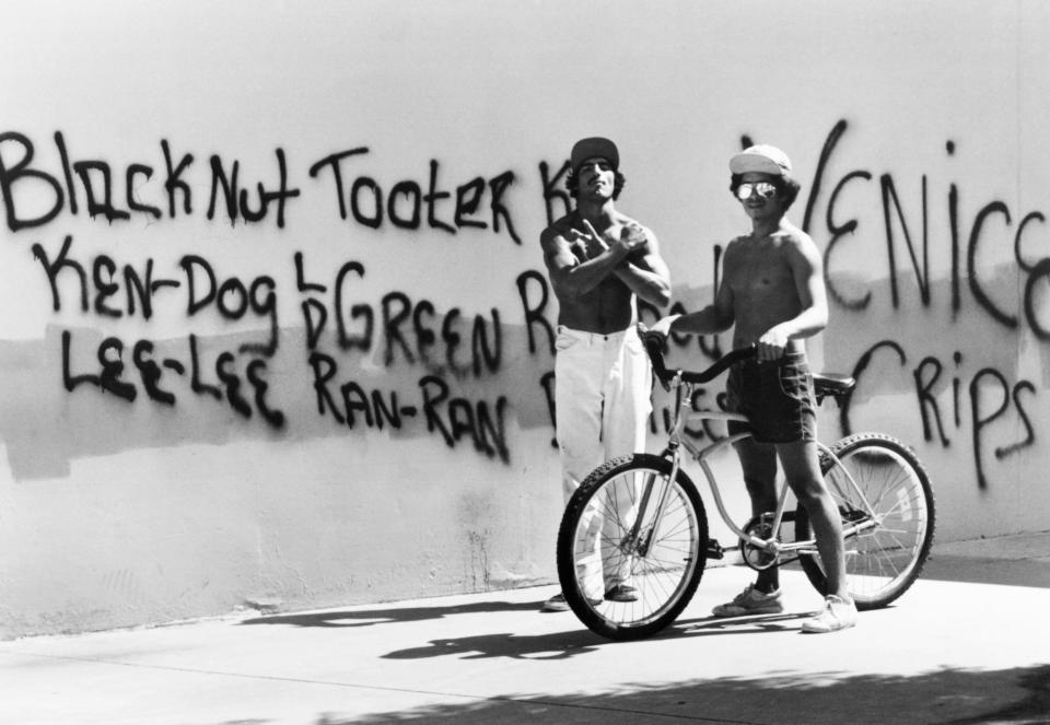  Two young men stand in front of a wall bearing the names of fellow gang members