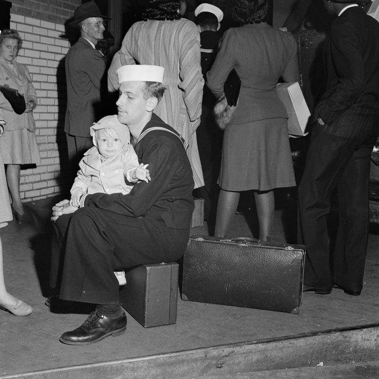  A sailor waits on the pavement with a baby, while waiting for a bus