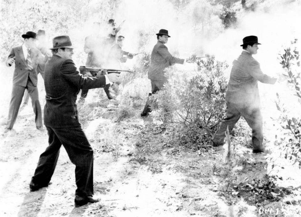  A group of gangsters wearing suits and hats practice firing their weapons in shrub land in the 1930s