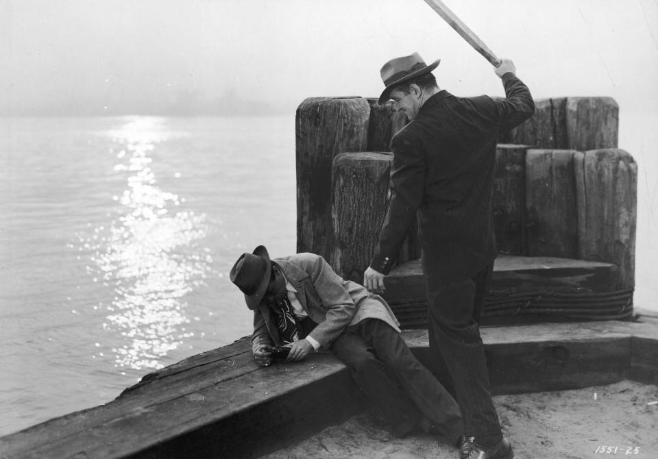  A man raises a stake to beat a drunken man sitting on a pier with a beer bottle