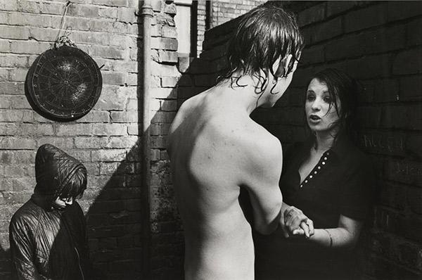  ‘Young couple in a backyard, 1975’. During a waterfight in a backyard a young woman gives a talking-to to her boyfriend. A child stands in the background with hood up to cover her hair