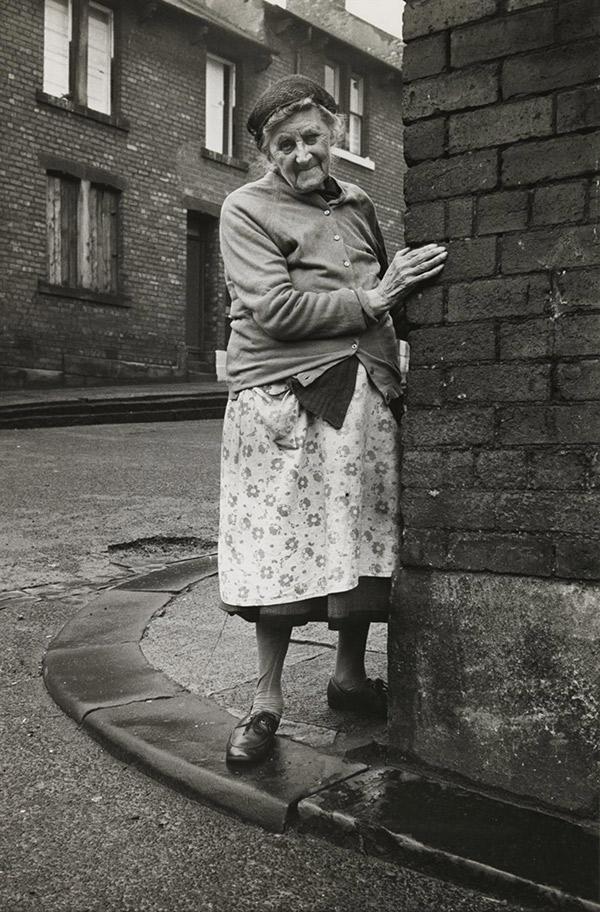  ‘Mrs Potter in Mason Street, 1975'. An elderly lady stands at the corner of a street wearing a floral apron, buttoned-up cardigan and a hairnet