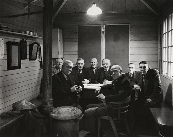  ‘Byker Park Dominoes Club, 1974’. A group of elderly men are pictured playing dominos in a wood-panelled hut. The gentlemen all face the camera and are smartly dressed in suits and ties