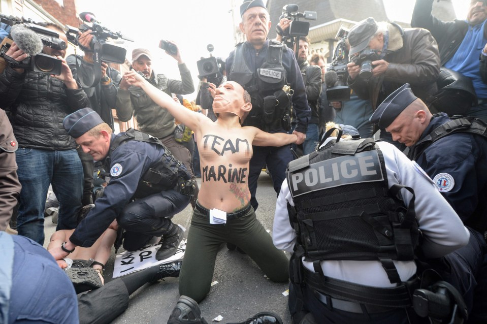 A masked demonstrator grabs press and police attention outside far right leader Le Pen’s local polling station