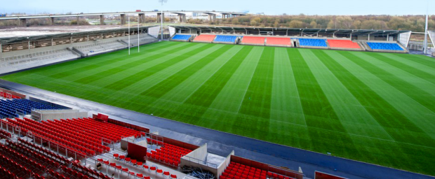 a soccer field with red and blue seats and a bridge in the background