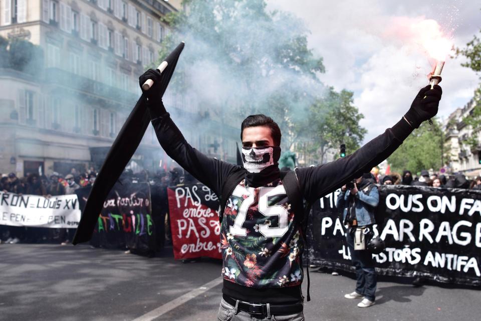  A man in a skeleton mask holds a black anarchist flag and a flare aloft as he leads the May Day march towards police lines