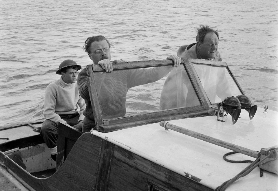 Sean sits in the back of a small boat piloted by John Mills as Richard Attenborough stands alongside him