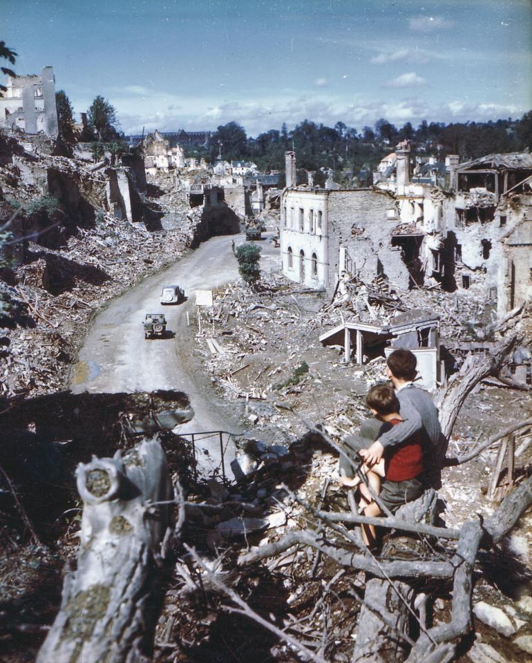  Two children survey the damage from the ruins of a castle rampart as American Jeeps make their way through Saint-Lo