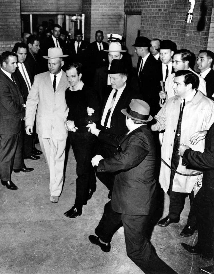 Lee Harvey Oswald is escorted to the Dallas city jail as nightclub owner Jack Ruby lunches with his gun