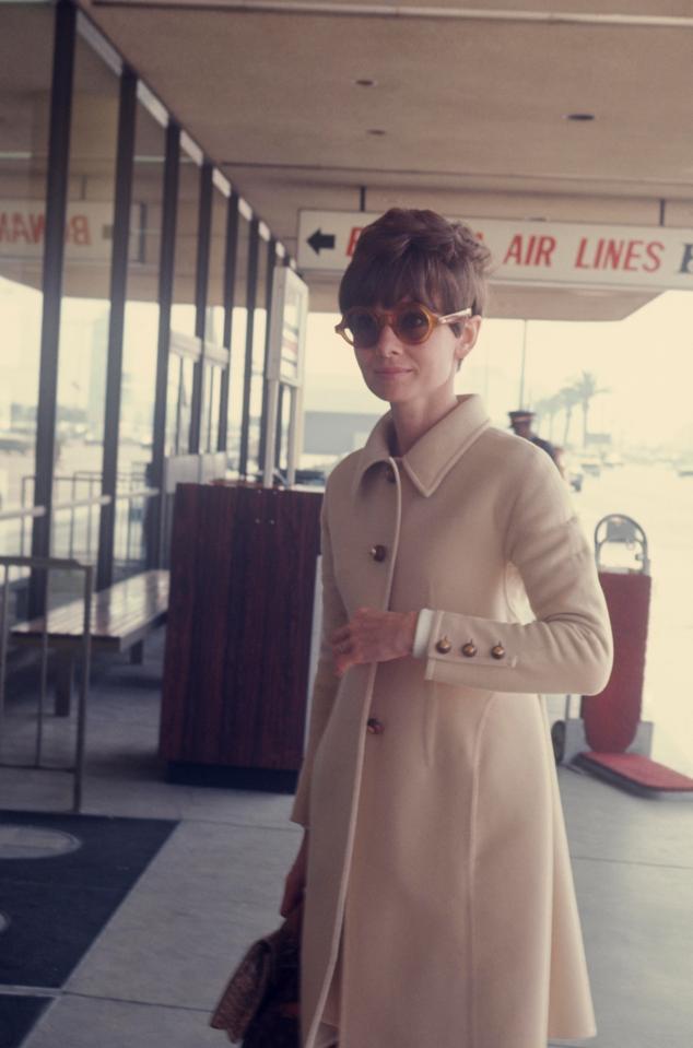  1970: Audrey is caught off guard at an airport in New York, wearing a classic cream coat and bold sunglasses