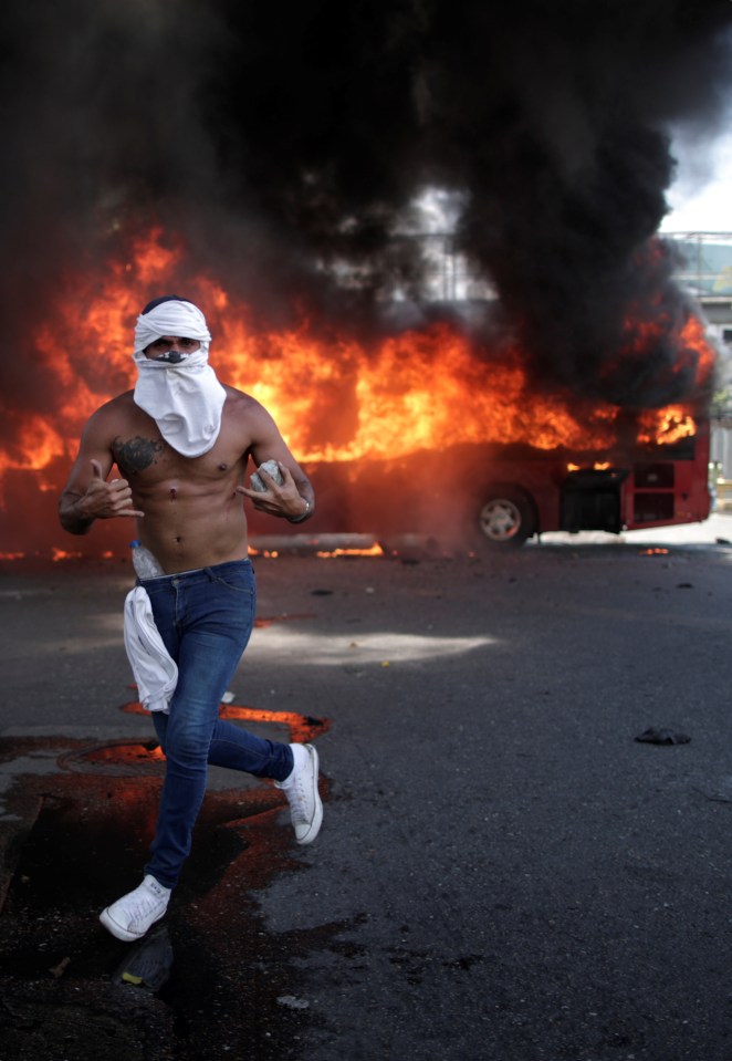  An opposition demonstrator gestures in front of a burning bus, while holding a rock