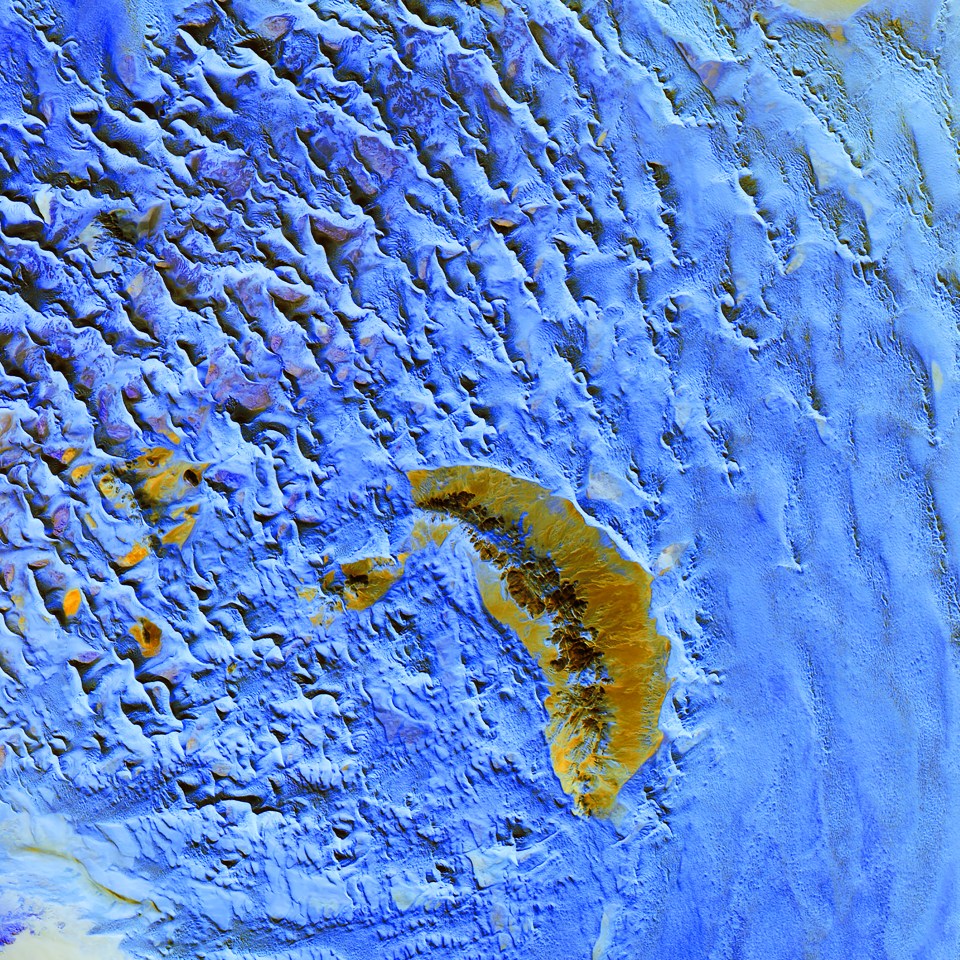  Resembling choppy waters in a rough sea, this image is an infrared exposition of a sand sea in Namibia. Sand dunes surround a crescent-shaped rocky hill. This inselberg is hard rock that resisted the erosion that took place over time around it. The inselberg disrupts the deposition of sand, changing the dune pattern.