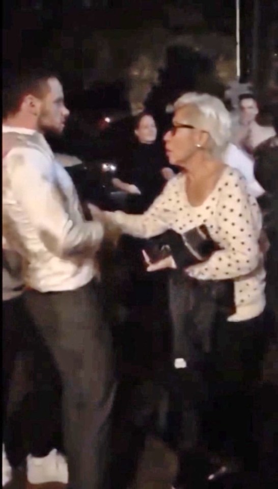 An elderly woman stands between Liam and the doorway