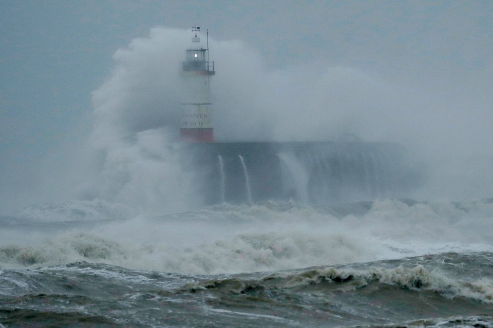 Waves crash over the harbour and a lighthouse in Newhaven