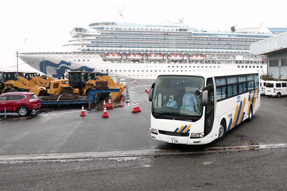  A bus with a driver wearing protective gear departs from the dockside next to the Diamond Princess cruise ship