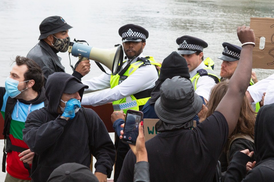 An officer holds his hands out as a man with a megaphone speaks on the bridge