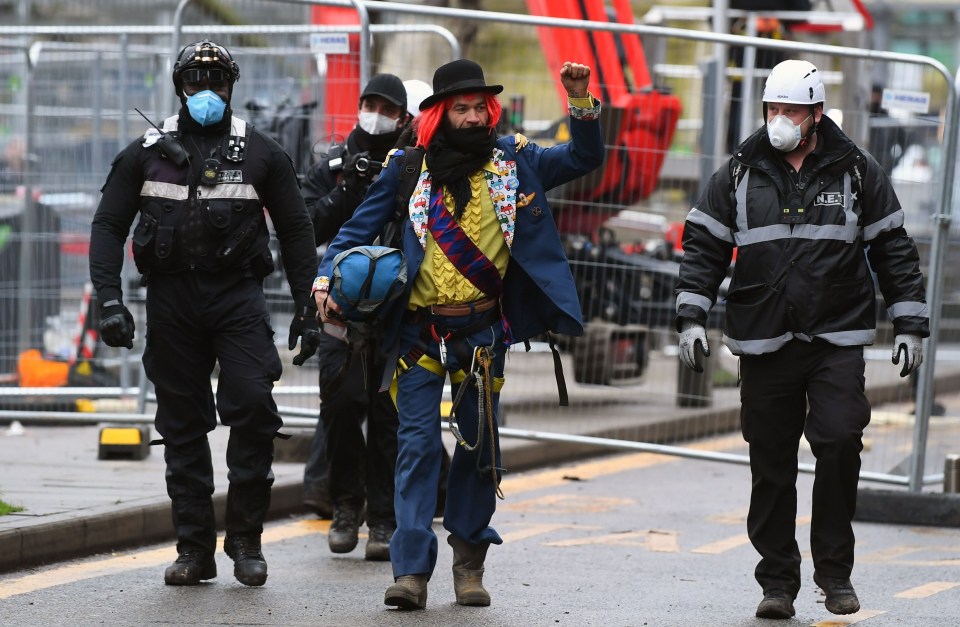 A brightly dressed man waves his fist as he is lead away