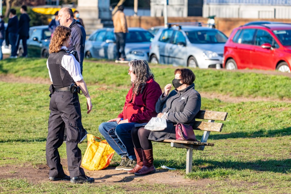 A police officer speaks to two people sitting on a bench on Wimbledon Common in London