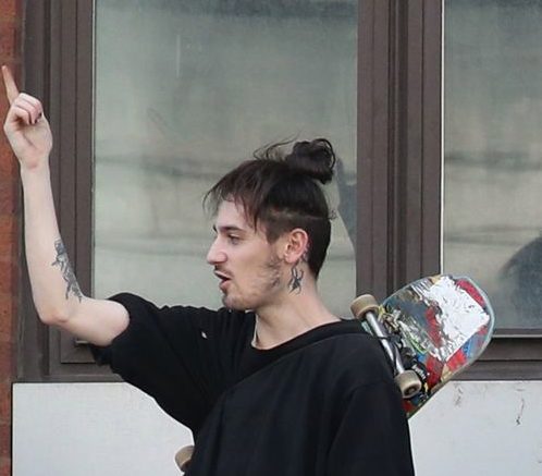 A protester with a top knot and neck tattoo carries a skateboard on his back