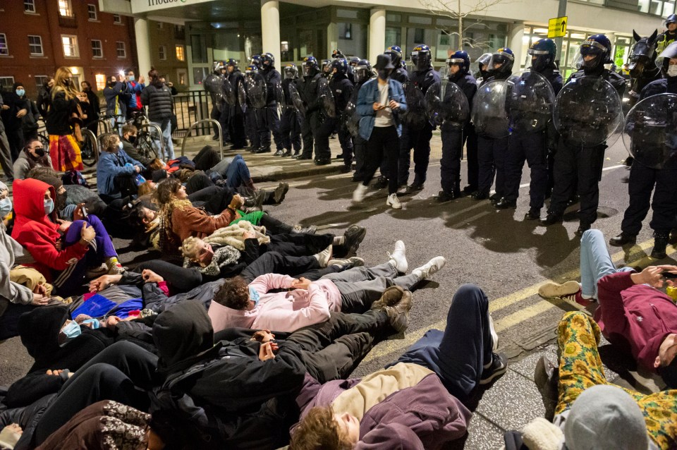 A crowd kneel and sit on the road in front of a line of riot police