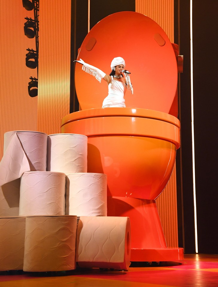 Katy pops out of a giant sized toilet, complete with enormous loo rolls