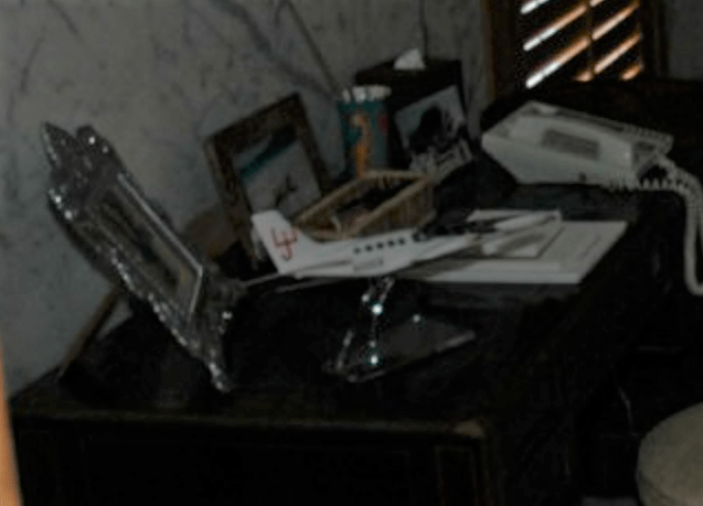 A framed nude photo of Ghislaine Maxwell sits on a desk near a small model plane and a telephone