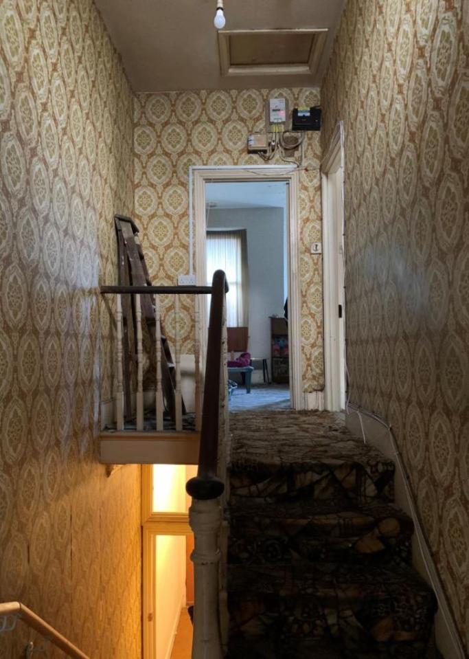 A hallway plastered in ancient, beige wallpaper and fuzzy carpet with a bannister in need of replacement