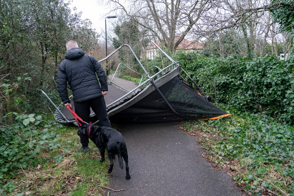 A dog walker navigates a trampoline swept out of a garden in East London today