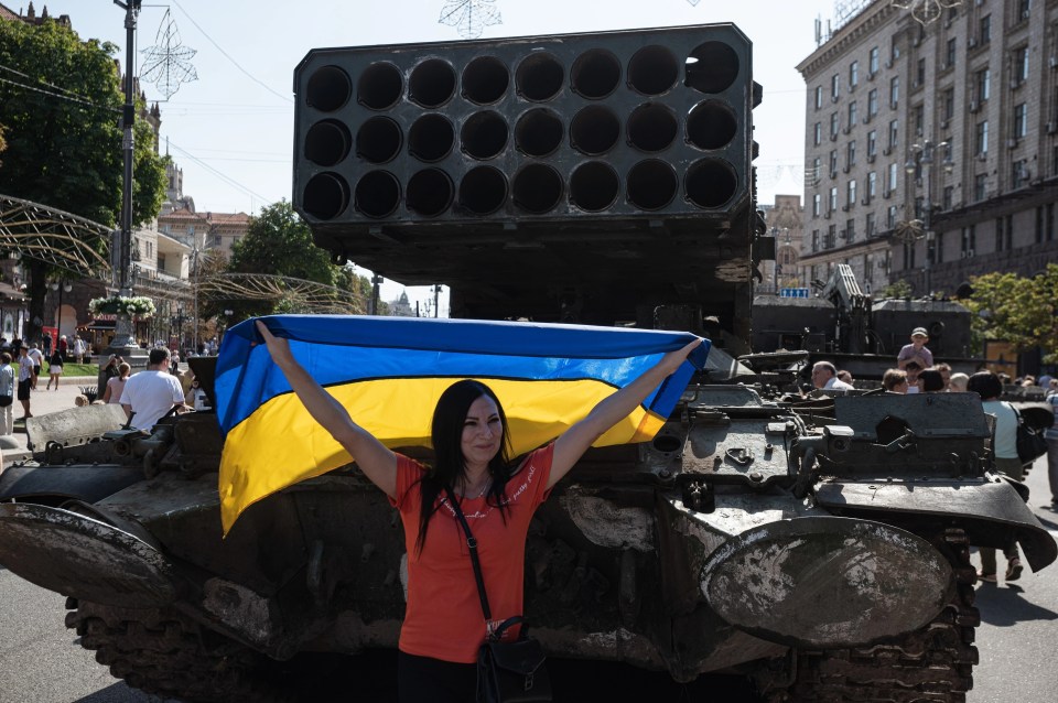 A woman holds a Ukrainian national flag as she poses in front of a Russian thermobaric rocket launcher