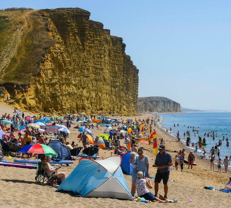 The beach is busy with holidaymakers and sunbathers enjoying the scorching hot sunshine and clear blue skies at the seaside resort of West Bay in Dorset