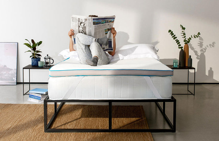 Person relaxing on a mattress topper while reading a newspaper.