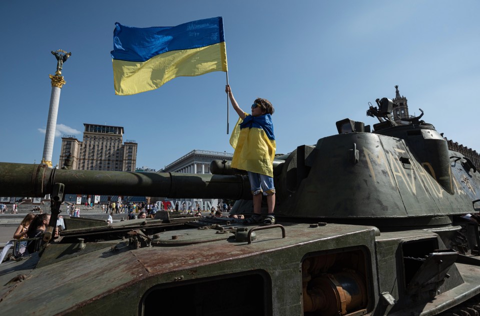 A boy wrapped in Ukrainian national flag holds a Ukrainian national flag as he stands on top of a Russian military vehicle