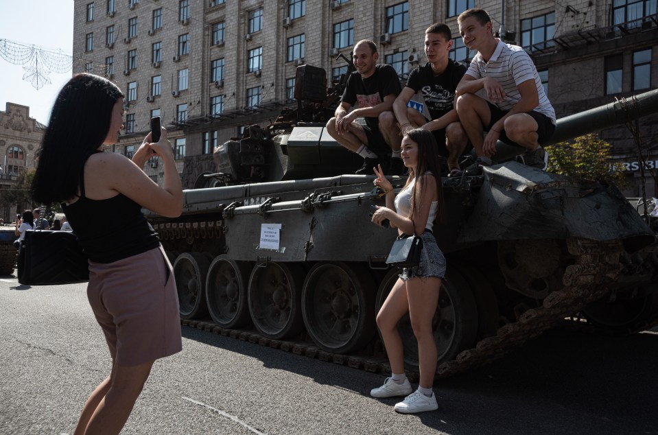 A young woman photographs her friends who pose for a picture by and on top of burnt tank