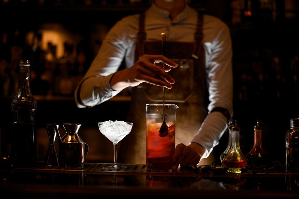 A male bartender in a white shirt and leather apron is stirring an alcoholic cocktail with a special bar spoon