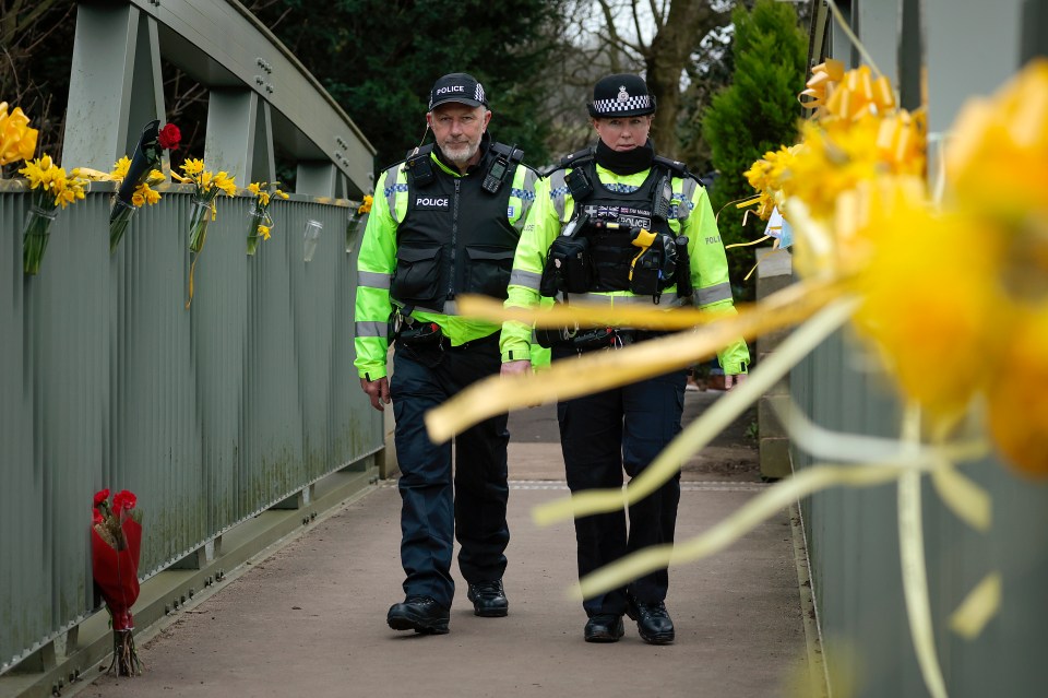 Police walk over a footbridge adorned with yellow ribbons and flowers