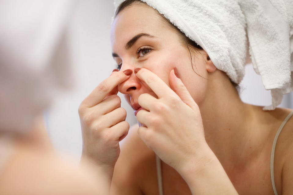 Reflection of woman in bathroom mirror wearing towel on hair after shower, squeezing blackead on her nose side with index fingernails