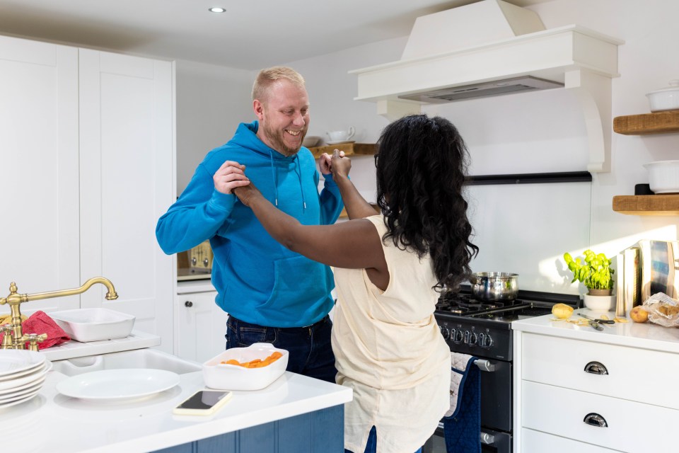 Mid-adult male and female married couple dancing in their kitchen smiling at each other. They are both wearing casual clothing.