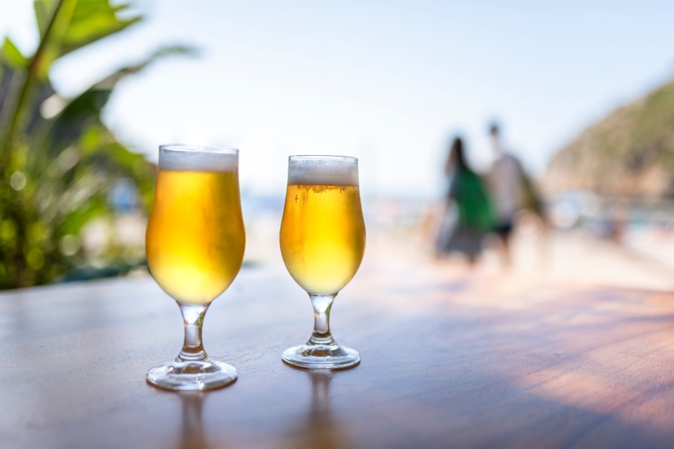 Two glasses of beer, with two people out of focus and the sea in the background