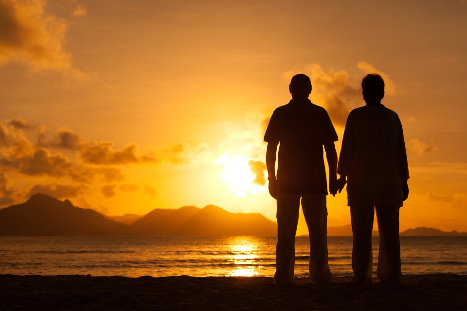 back view silhouette couple standing together on beach watching sunset over island