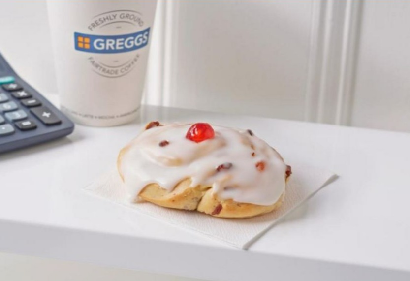 a cinnamon roll with a cherry on top sits on a napkin next to a cup of greggs coffee