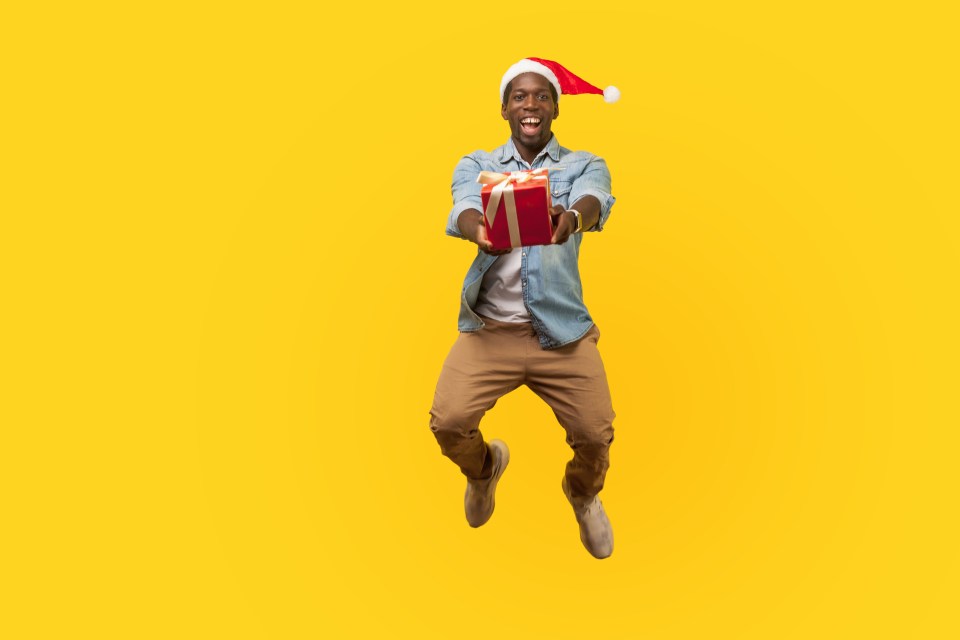 Full length portrait of extremely happy young man in santa hat and casual denim shirt jumping or flying and giving christmas gift box to camera. indoor studio shot isolated on yellow background; Shutterstock ID 1532807102; purchase_order: -; job: -; client: -; other: -