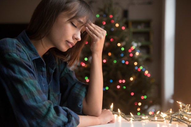 A young woman sits at a table, head in hand, looking sad in front of a Christmas tree.