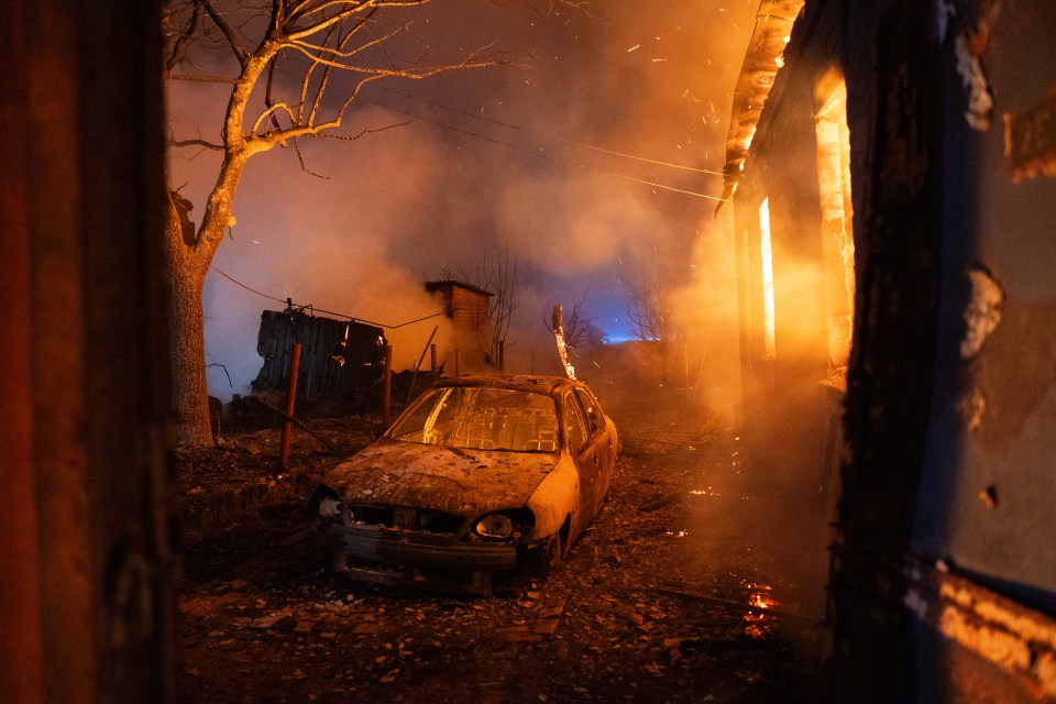 A burnt car stands near a burning private house