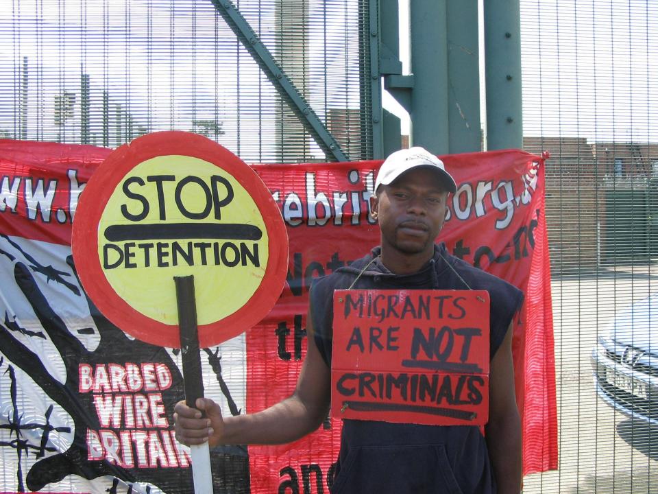 Anicet Mayela, wearing a 'Migrants are not criminals' sign protests at  Campsfield House Detention Centre in Kidlington, Oxford