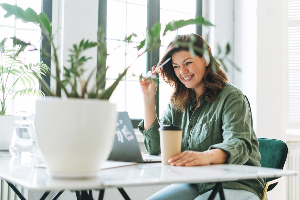 Young smiling brunette woman plus size working at laptop on table with house plant in bright modern office